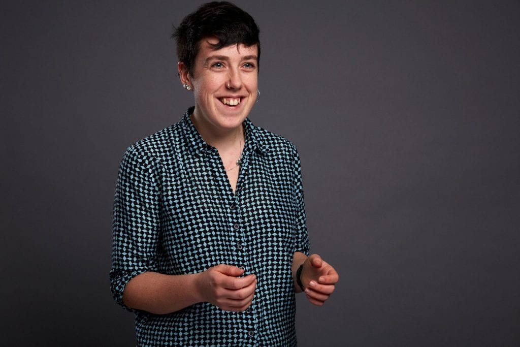 A photo of Jen Slater, a white person with short brown hair, on a grey background. Jen is wearing a blue and black checked shirt. They have three earrings in their left ear (which is on show), a nose ring, and a thin silver necklace. They are looking just off centre of the camera, smiling and gesturing with their hands, as if mid-sentence.