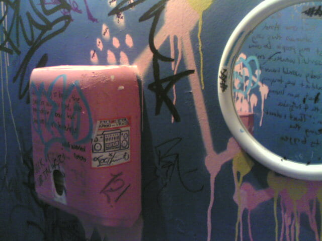 Toilet Wall showing hand dryer and mirror. Dark blue wall and hand dryer (painted poorly in pink) covered in graffiti including reflection in mirror.