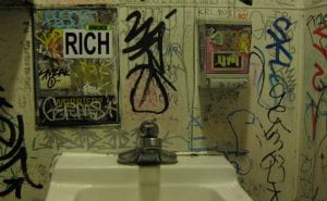 White ceramic sink. The wall it is mounted on is covered in graffiti and posters, mostly tags and writing.