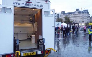 A mobile changing places toilets visible in back of a van, at an event in a central London square. The weather looks rainy and cold.
